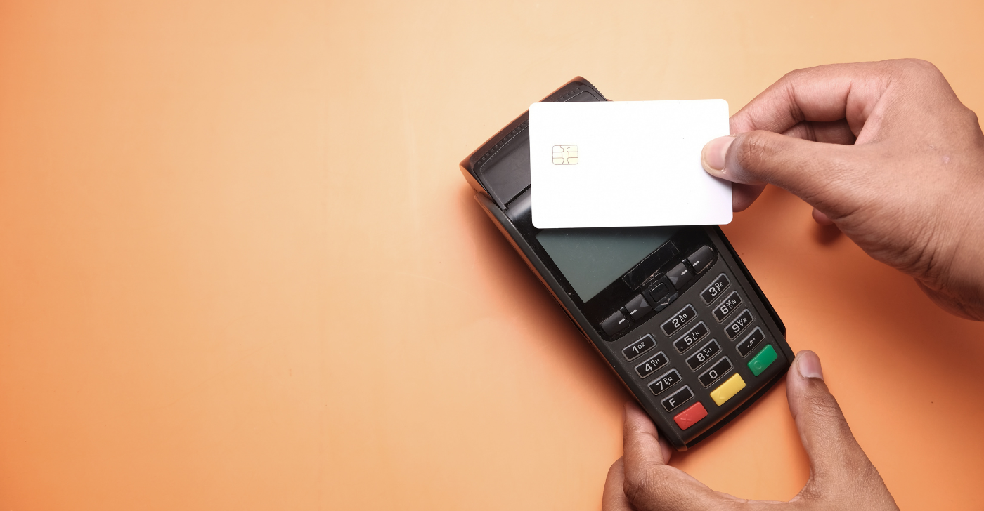 A black EFTPOS machine on an orange background with a white blank bank card held in front of it.