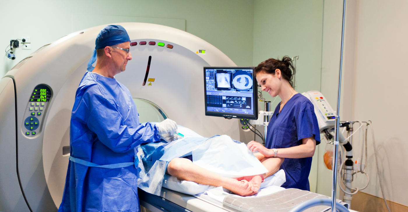 A white male medical professional stands opposite a white female medical professional. They are in either side of a patient about to enter an MRI machine.
