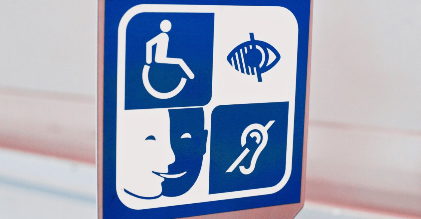 White-blue sign that features four accessibility symbols: a wheelchair symbol, a visual impairment symbol, the S3A pictogram and a hearing loop symbol.