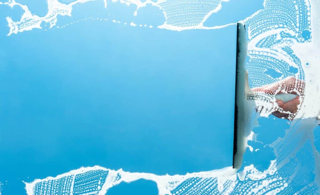 A photo of a hand holding a window wiper and cleaning a window covered with foam. The photo is intended to allude to content spring cleaning, which is the topic of the article.