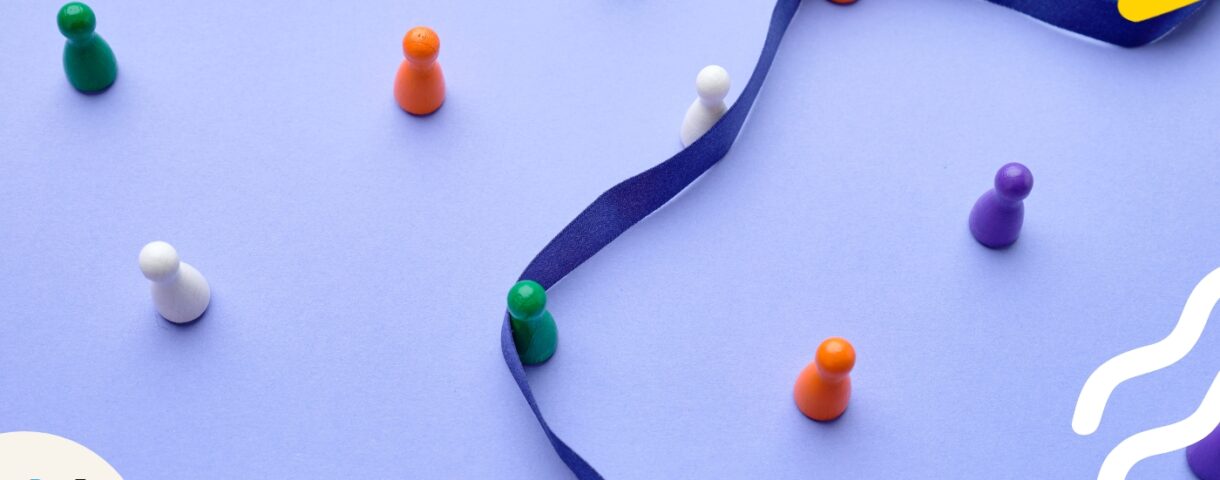 Ein blaues Satin-Band dient als Orientierung für bunte Holzfiguren und steht symbolisch für das Schreiben nach Vorgaben.